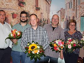 Sechs Personen stehen nebeneinander vor einer Wand mit mediterranem Wandbild, jede hält einen Blumenstrauß mit verschiedenen Blumen wie Sonnenblumen und Rosen.
