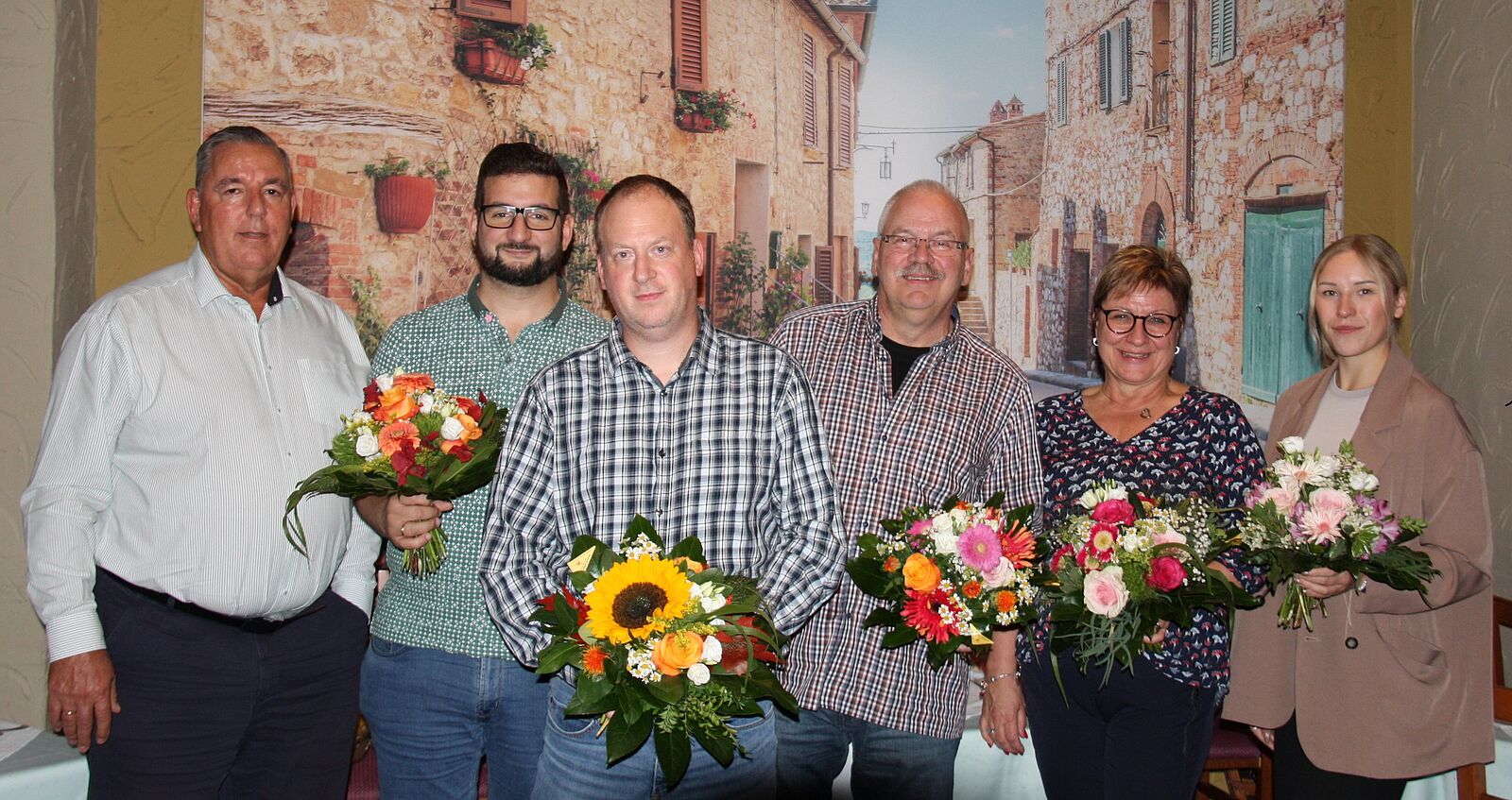 Sechs Personen stehen nebeneinander vor einer Wand mit mediterranem Wandbild, jede hält einen Blumenstrauß mit verschiedenen Blumen wie Sonnenblumen und Rosen.