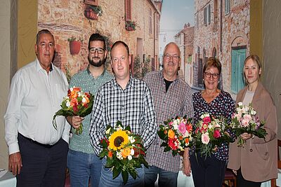 Sechs Personen stehen nebeneinander vor einer Wand mit mediterranem Wandbild, jede hält einen Blumenstrauß mit verschiedenen Blumen wie Sonnenblumen und Rosen.
